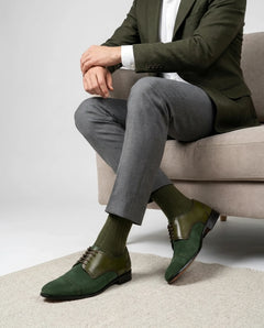 Lifestyle image of HAC’s Plush Lace Up oxford shoes styled with formal outfit for a refined menswear look. Person wearing a green suit with gray pants and green shoes sitting on a beige couch.